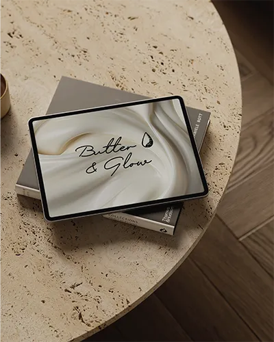 Tablet showing the Butter & Glow logo on a creamy swirl background, resting on books on a travertine table