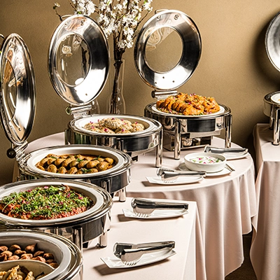 A catering buffet of open chafing dishes with hot dishes, set on white tablecloths with blossom branches behind