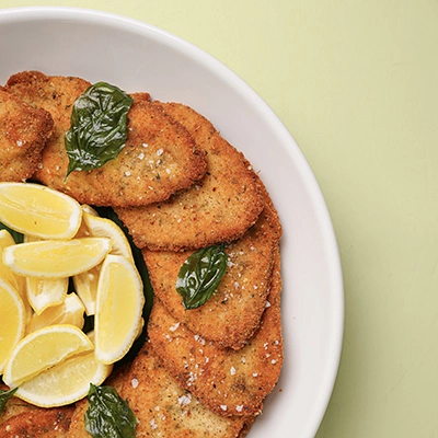Golden breaded cutlets with lemon wedges and fresh basil in a white bowl on a pale green background